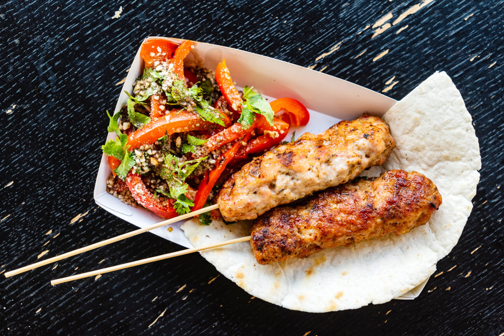 Two grilled chicken kabobs are served on a piece of flatbread with a side salad of mixed greens and red peppers, all presented in a white takeout container on a dark wooden surface.