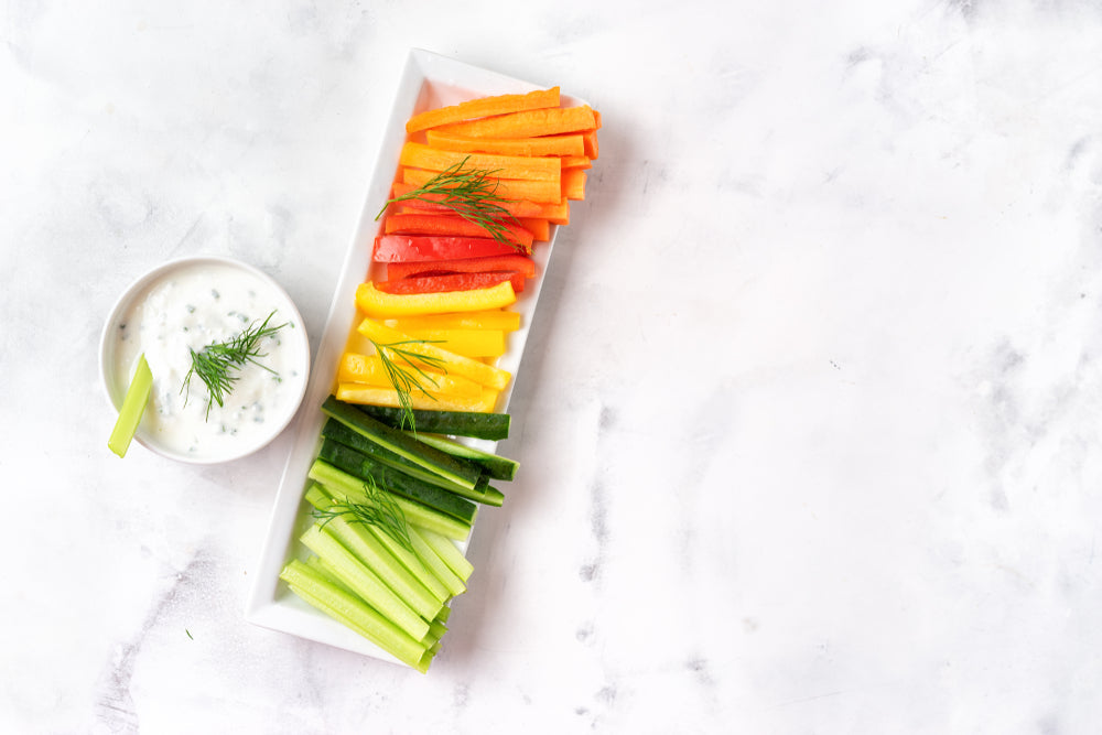 A white platter holds colorful strips of carrots, red and yellow bell peppers, cucumbers, and celery arranged in a rainbow pattern, alongside a small bowl of creamy dip garnished with dill.