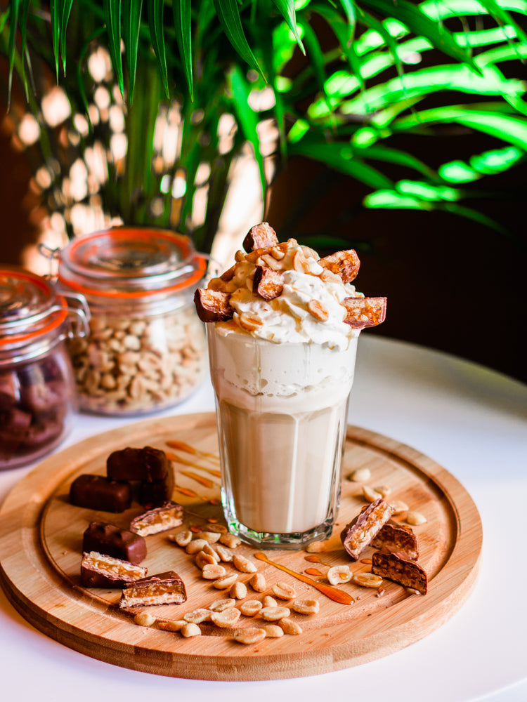 A tall glass filled with a creamy chocolate peanut butter shake topped with whipped cream and chopped candy sits on a wooden platter surrounded by peanuts and chocolate bars, with green foliage in the background.