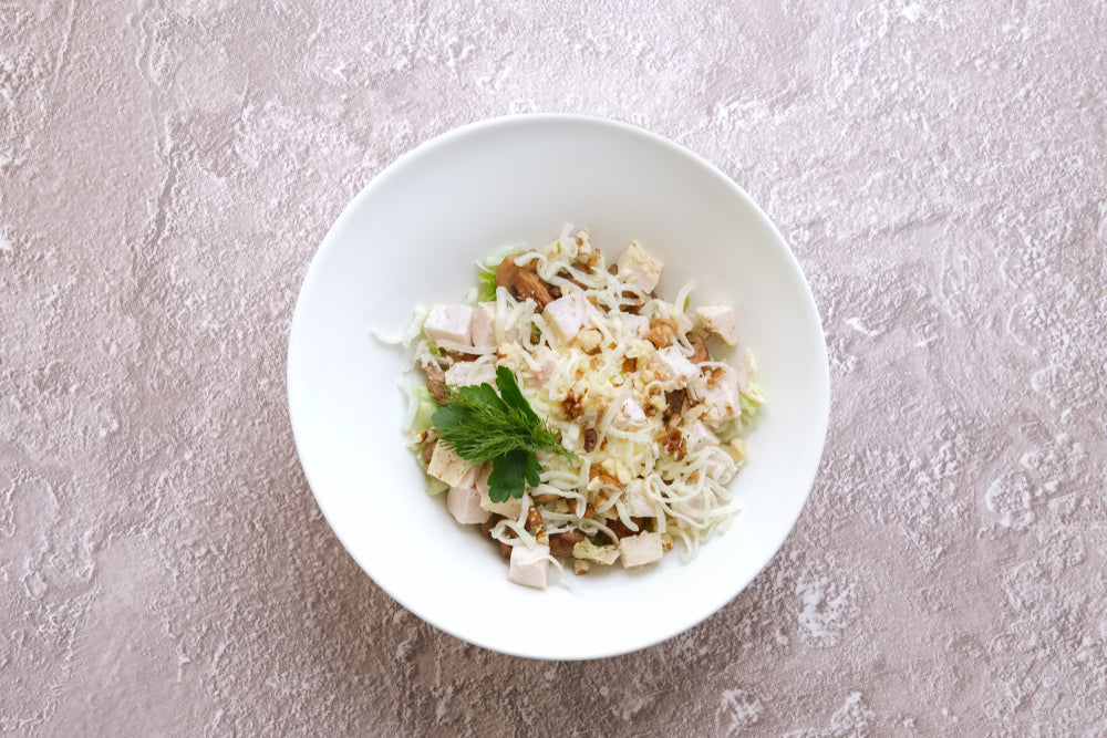 A white bowl contains a salad with shredded cabbage, diced chicken, nuts, and a sprig of mint, set against a textured light pink background.