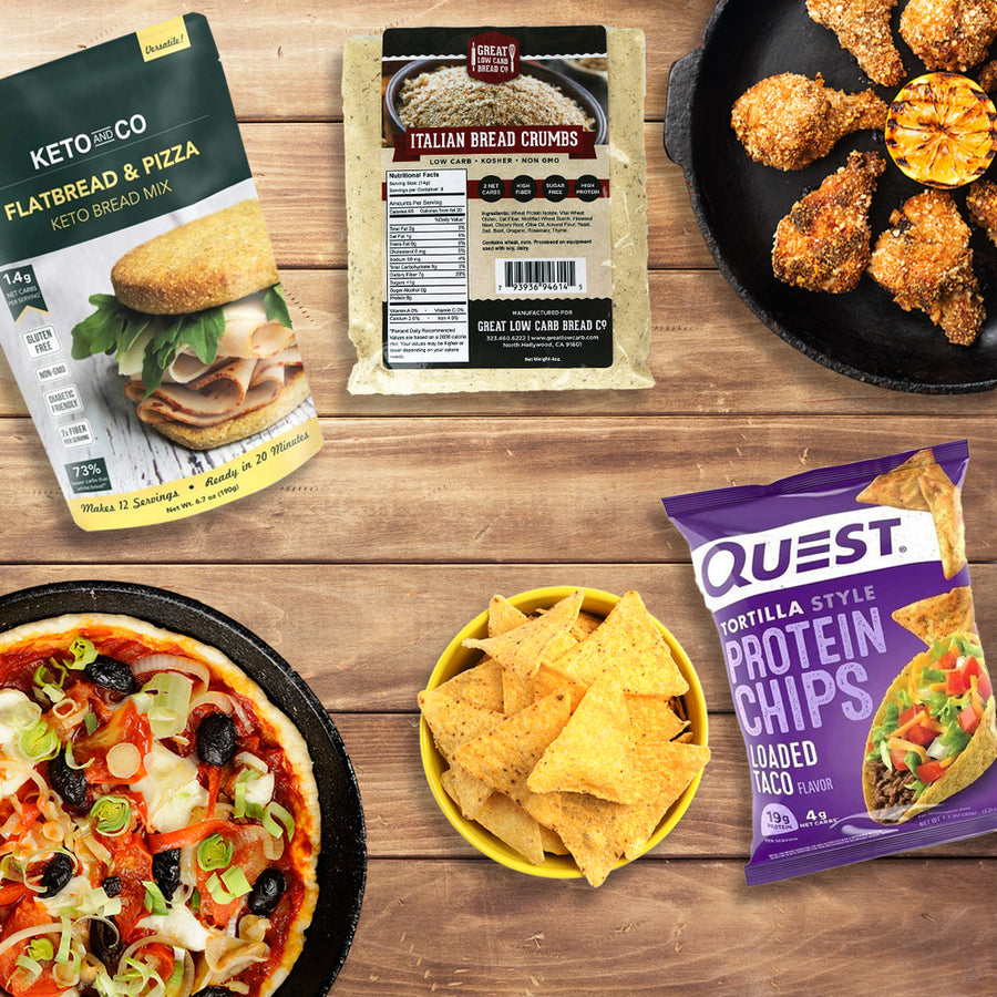 A variety of bariatric-friendly snacks for game day is displayed, including a keto flatbread mix, Italian bread crumbs, protein chips, and a pizza topped with vegetables and olives, all arranged on a wooden table.
