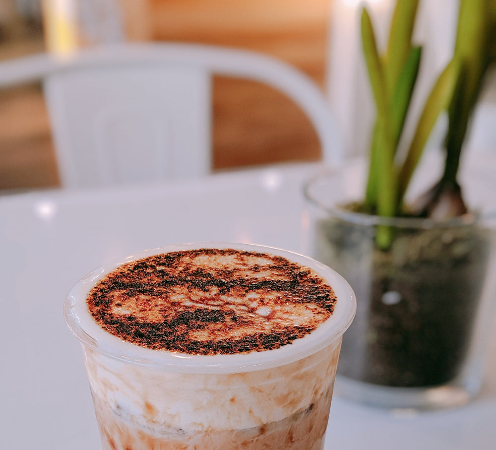 A clear cup filled with a creamy, layered shake topped with toasted marshmallow and chocolate sprinkles sits on a table, with a small potted plant blurred in the background.