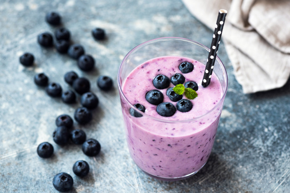 A glass of purple-blue smoothie topped with fresh blueberries and a mint leaf sits on a textured surface, with scattered blueberries nearby and a striped straw in the drink.