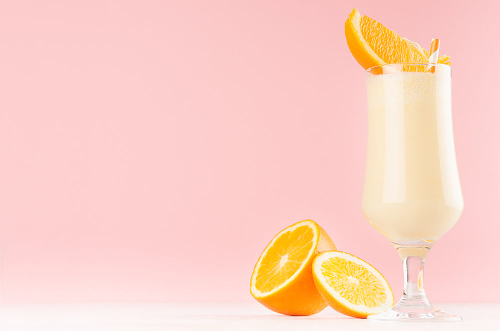 A tall glass of creamy orange shake, garnished with orange slices, sits beside whole and halved oranges against a soft pink background.