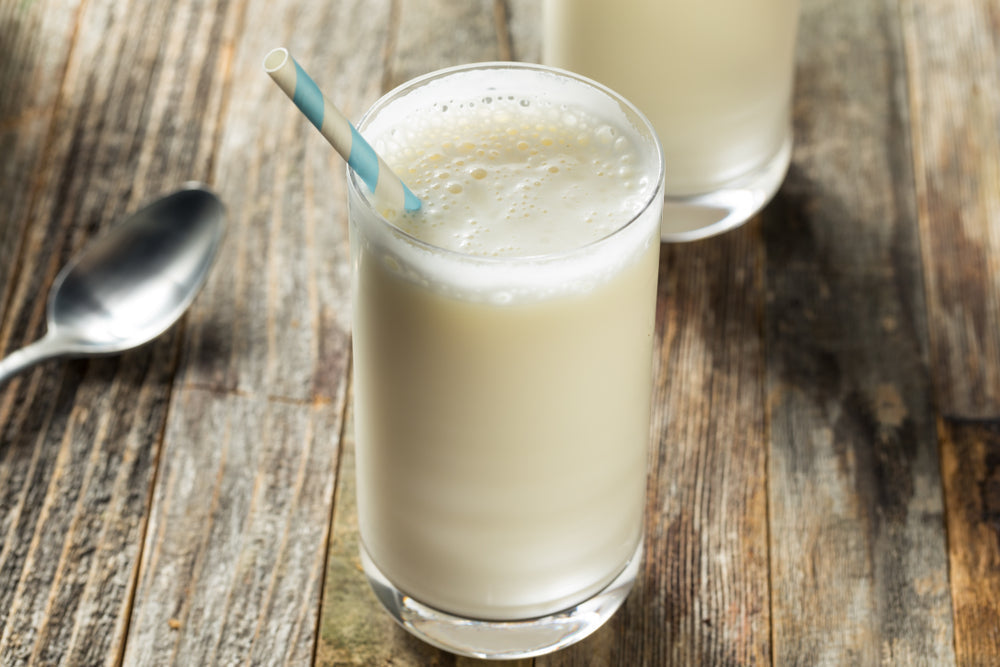 Two tall glasses of creamy peppermint shake with a light blue and white striped straw sit on a rustic wooden table, with a silver spoon nearby.