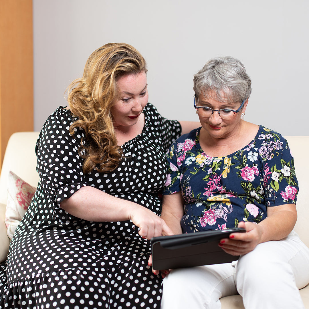 Two women sit closely on a couch, one in a polka dot dress pointing at a tablet while the other, wearing glasses and a floral top, looks attentively at the screen, discussing weight loss surgery.