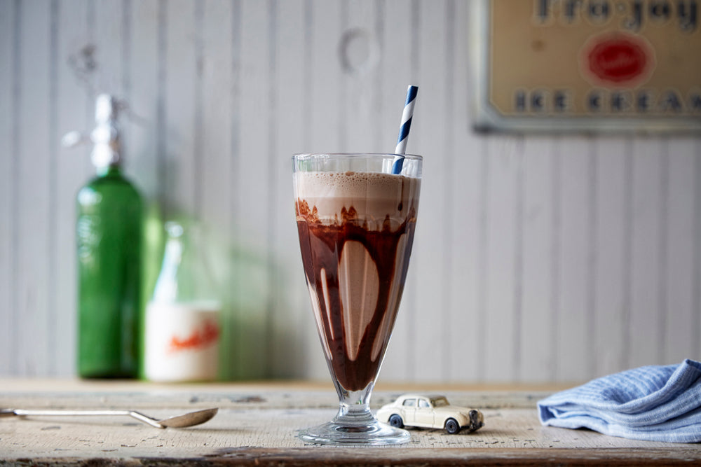A tall glass of chocolate ice cream soda with a blue striped straw sits on a wooden table, surrounded by a small toy car, a spoon, and vintage bottles in a softly lit setting.