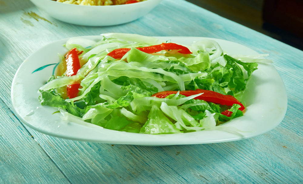 A white plate holds a fresh salad of shredded green lettuce and red bell pepper strips, placed on a light blue wooden table.