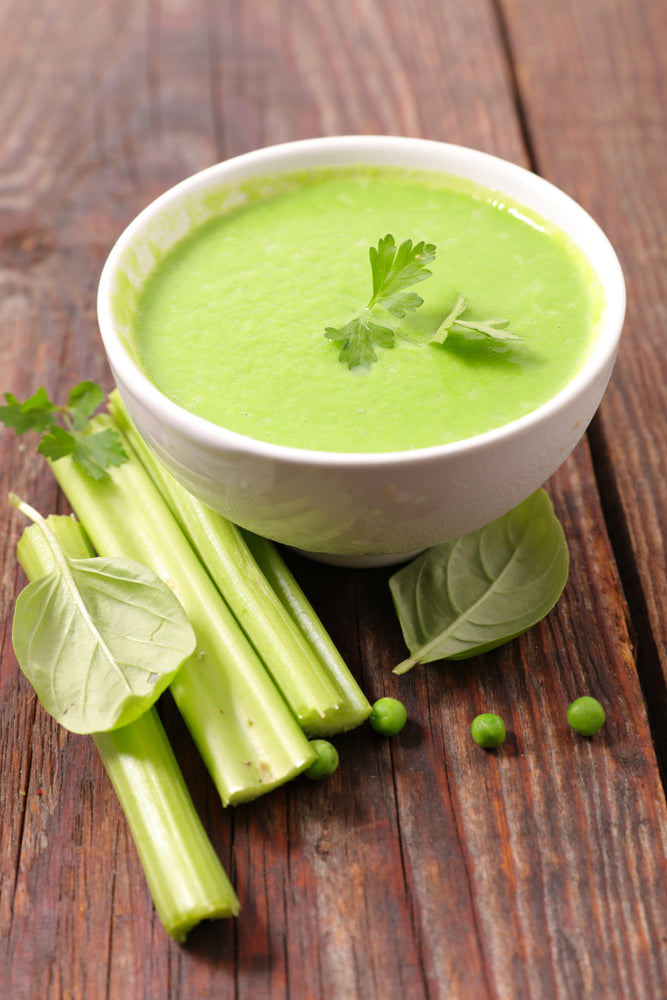 A bowl of vibrant green celery soup garnished with fresh herbs sits on a wooden table, accompanied by stalks of celery and green peas.