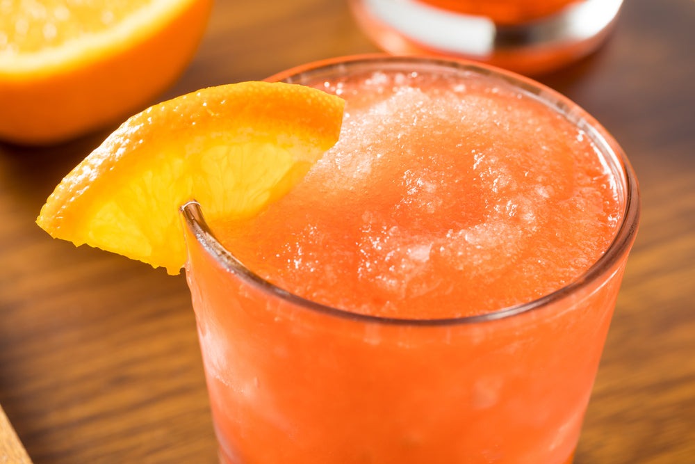 A frosty orange drink sits in a glass, topped with a slice of orange, on a wooden surface, with a blurred orange in the background, suggesting a refreshing summer beverage.