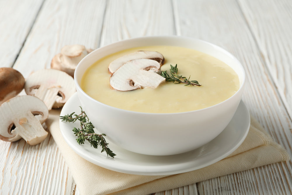 A creamy bowl of mushroom soup is garnished with sliced mushrooms and fresh thyme, placed on a light-colored napkin against a rustic wooden background.