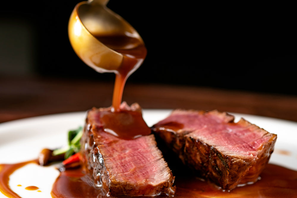 A spoon pours rich brown sauce over two slices of juicy, pink beef on a white plate, accompanied by colorful vegetables, set against a dark background.