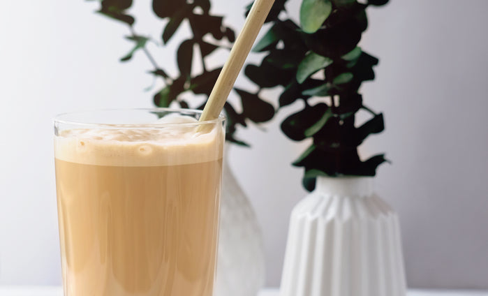 A glass of creamy French vanilla root beer float with a wooden straw sits in front of a white vase and green leaves, creating a refreshing and inviting atmosphere.
