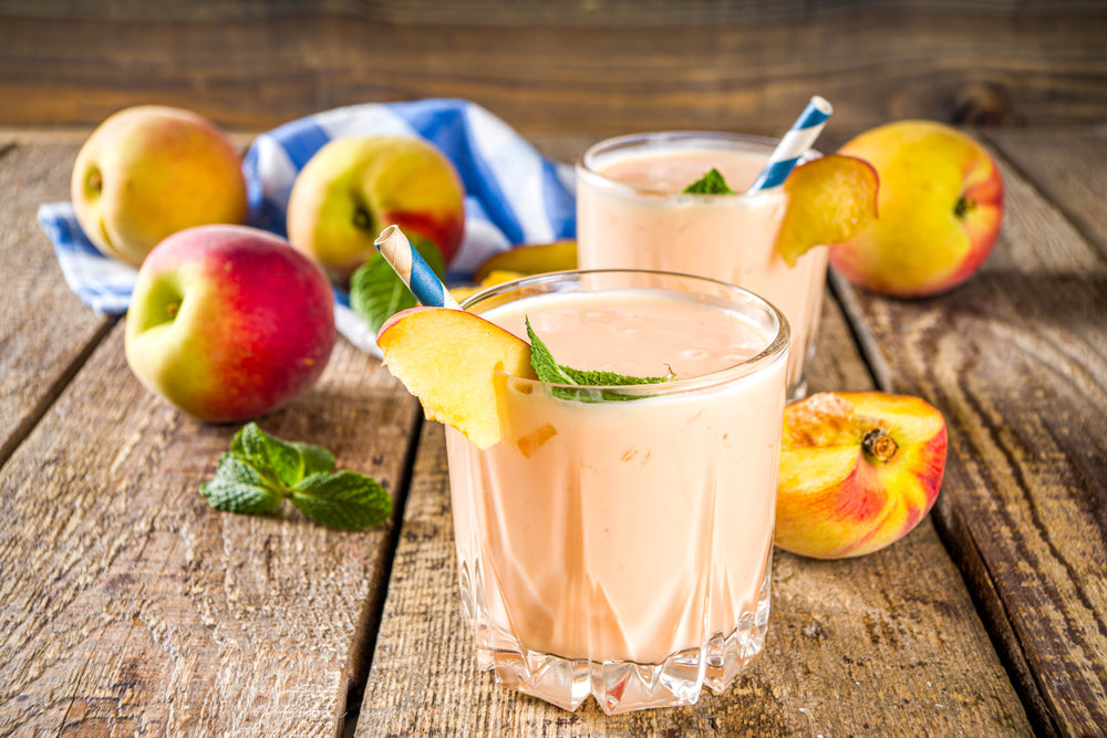 Two glasses of peach smoothie, garnished with peach slices and mint, sit on a wooden table surrounded by fresh peaches and a blue-striped cloth.