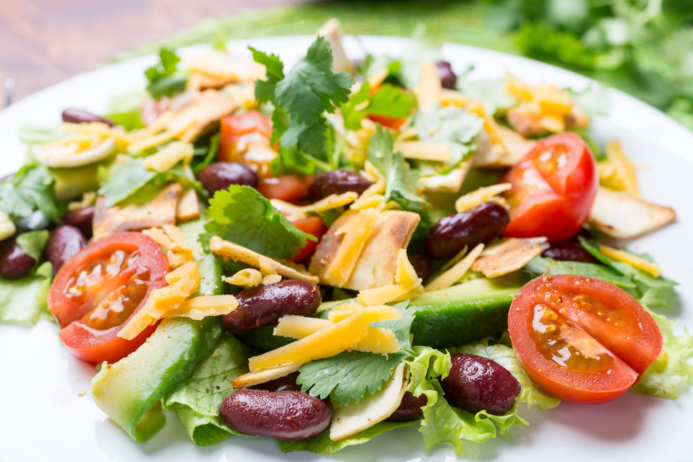 A vibrant Mexican salad features fresh lettuce, tomatoes, green bell peppers, kidney beans, tortilla strips, and shredded cheese, garnished with cilantro on a white plate.