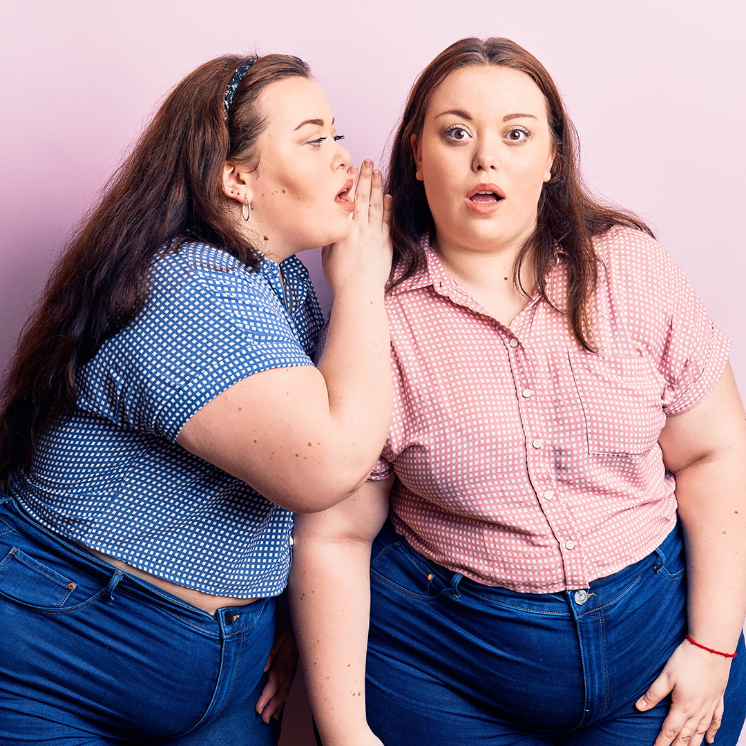 Two women stand side by side against a pink background; one leans in to whisper to the other, who looks surprised, both wearing patterned shirts and jeans, conveying a sense of secrecy and curiosity.