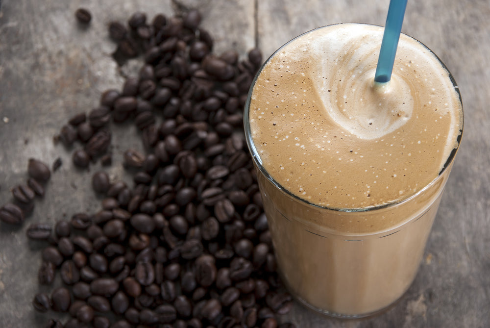 A tall glass of mocha vanilla shake with a blue straw sits on a wooden surface, surrounded by scattered dark coffee beans, showcasing a creamy, frothy top.