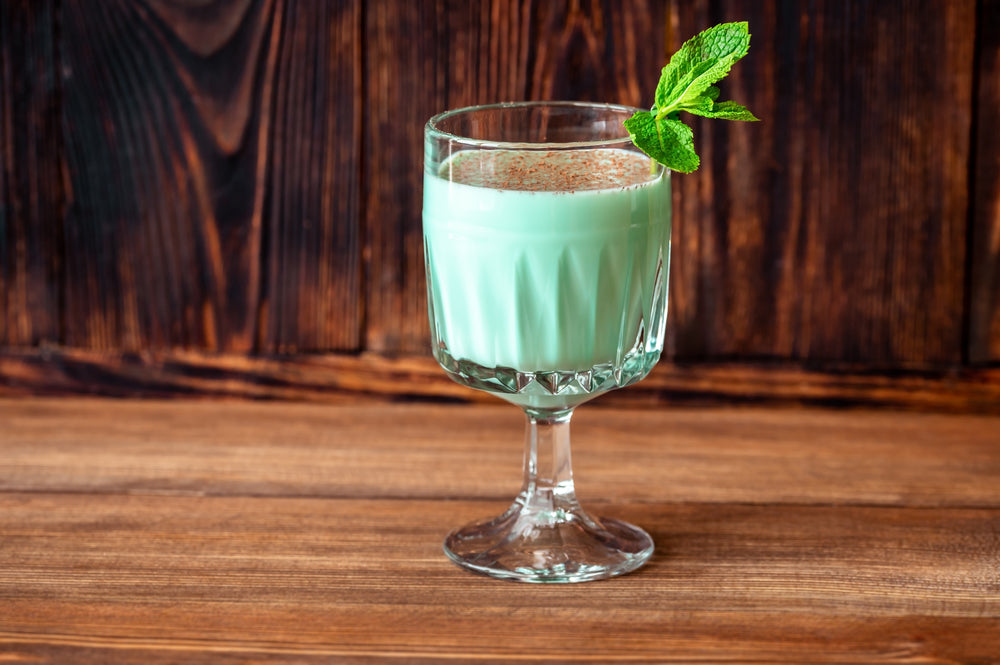 A glass of mint green Grasshopper Shake topped with a sprig of mint and a sprinkle of cocoa sits on a wooden table against a rustic wooden background.