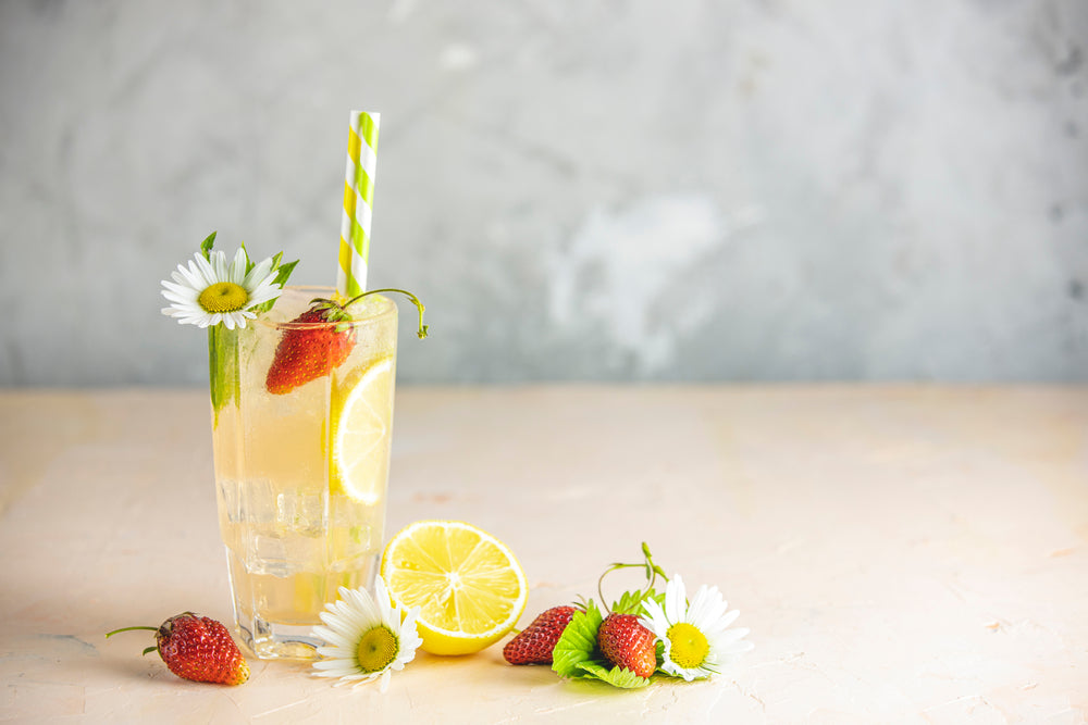 A tall glass of orange chamomile iced tea, garnished with lemon slices, strawberries, and daisies, sits on a light surface with a striped straw, creating a refreshing summer drink scene.