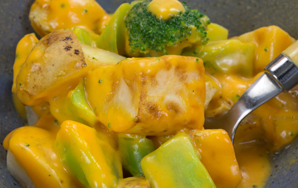A close-up of a bowl filled with diced potatoes, green bell peppers, and broccoli, all coated in a creamy orange cheese sauce, with a fork resting on the side.