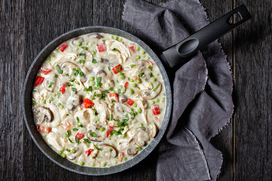 A black skillet filled with creamy A la King sauce, featuring chicken, mushrooms, red peppers, and green peas, sits on a wooden surface next to a folded gray cloth.