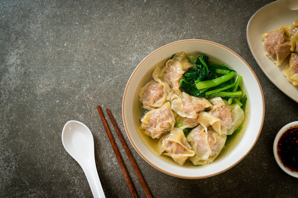A bowl of won ton soup filled with dumplings and leafy greens sits on a textured gray surface, accompanied by a small dish of chili sauce and chopsticks.
