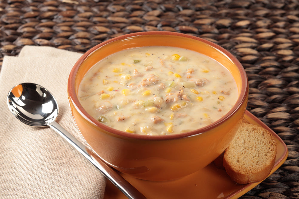 A warm bowl of creamy broccoli bisque with pieces of corn and herbs sits on a woven placemat, accompanied by a small slice of bread and a shiny spoon resting beside a folded napkin.