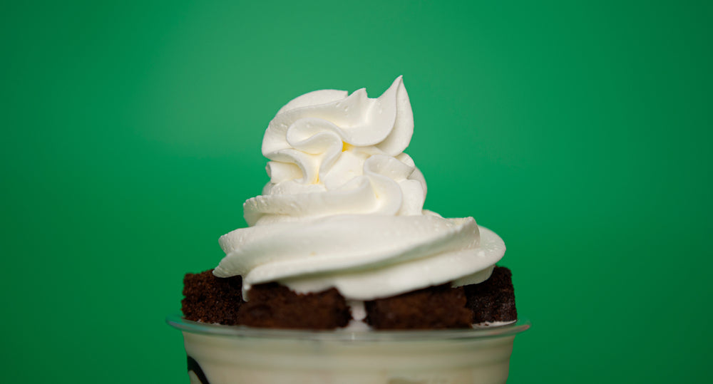 A close-up of a twisted cocoa smoothie topped with whipped cream and chocolate cake pieces, set against a bright green background.