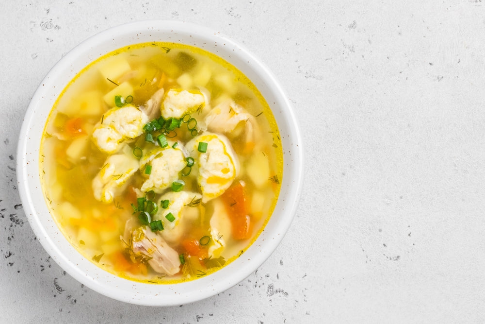 A white bowl filled with chicken and dumplings features tender chicken pieces, fluffy dumplings, and colorful vegetables in a savory broth, garnished with chopped green onions.