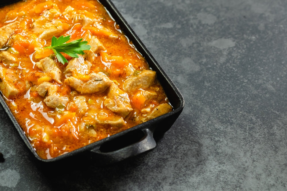 A black skillet holds a savory chicken gravy with tender pieces of chicken and chopped vegetables, garnished with a sprig of parsley, set against a dark, textured background.