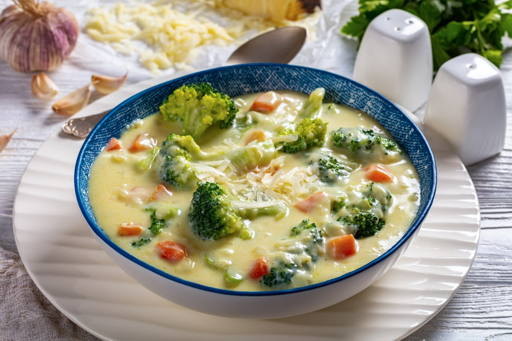 A blue bowl filled with creamy chicken and broccoli soup, featuring chunks of broccoli and carrots, sits on a white plate with garlic and cheese in the background, alongside salt and pepper shakers.