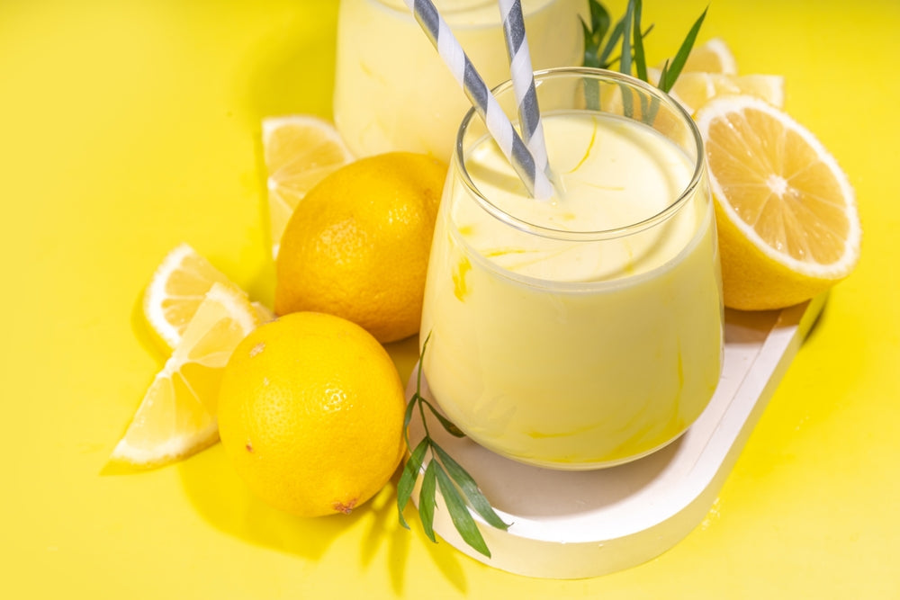 Two glasses of lemon shake with striped straws are placed on a white tray surrounded by whole and sliced lemons, set against a bright yellow background.