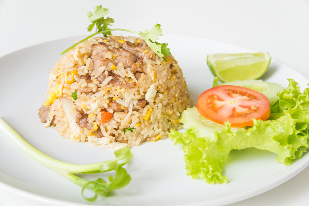 A plate features a mound of fried rice with bits of meat, garnished with green onions and cilantro, accompanied by a slice of lime, a tomato slice, and a serving of lettuce.