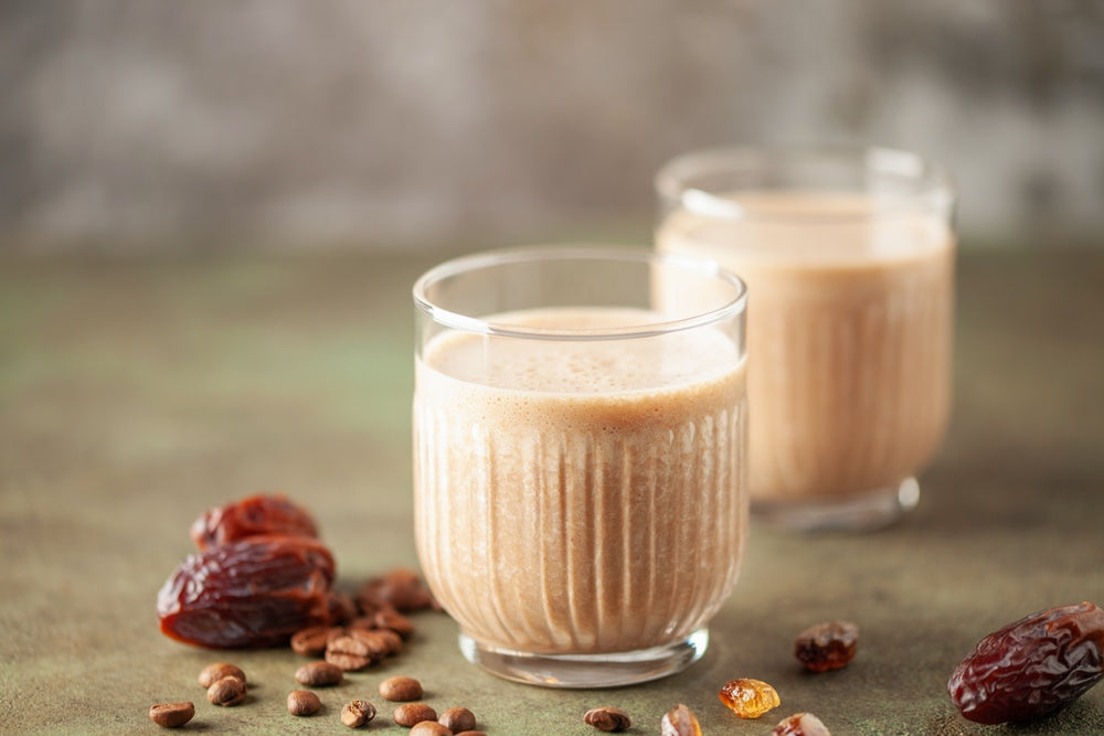 Two glasses of a creamy, brown shake sit on a textured surface, surrounded by dates and coffee beans, suggesting a rich and indulgent beverage.