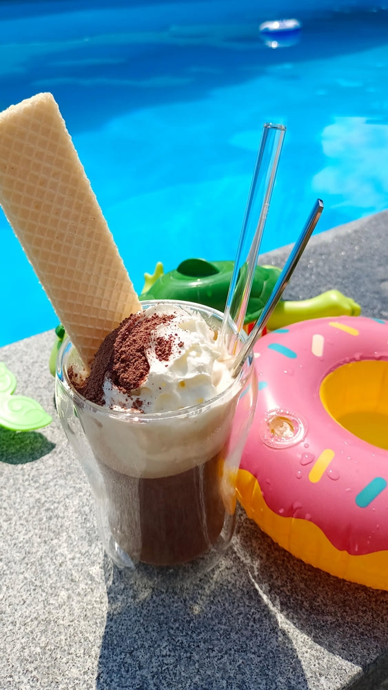 A tropical coffee shake topped with whipped cream and cocoa sits on a stone surface beside a colorful inflatable donut and a green toy, with a bright blue pool in the background.