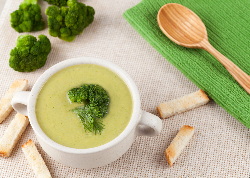 A bowl of creamy broccoli soup garnished with a small piece of broccoli and dill sits on a textured surface, accompanied by toasted bread pieces and a green cloth, with fresh broccoli in the background.