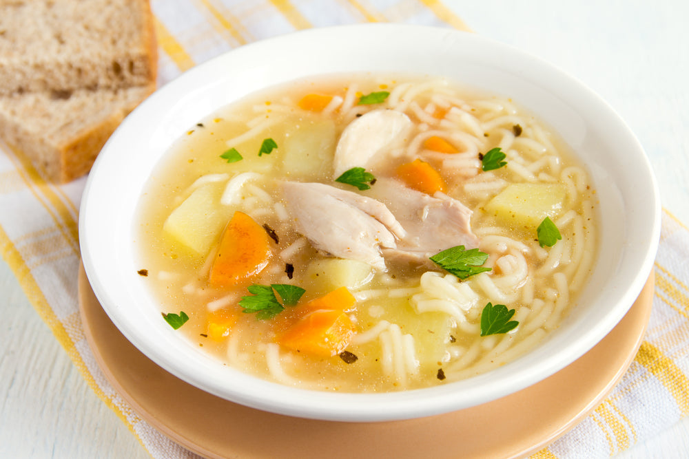 A bowl of chicken noodle soup with tender chicken pieces, carrots, and potatoes is garnished with parsley, accompanied by a slice of bread on a striped towel.