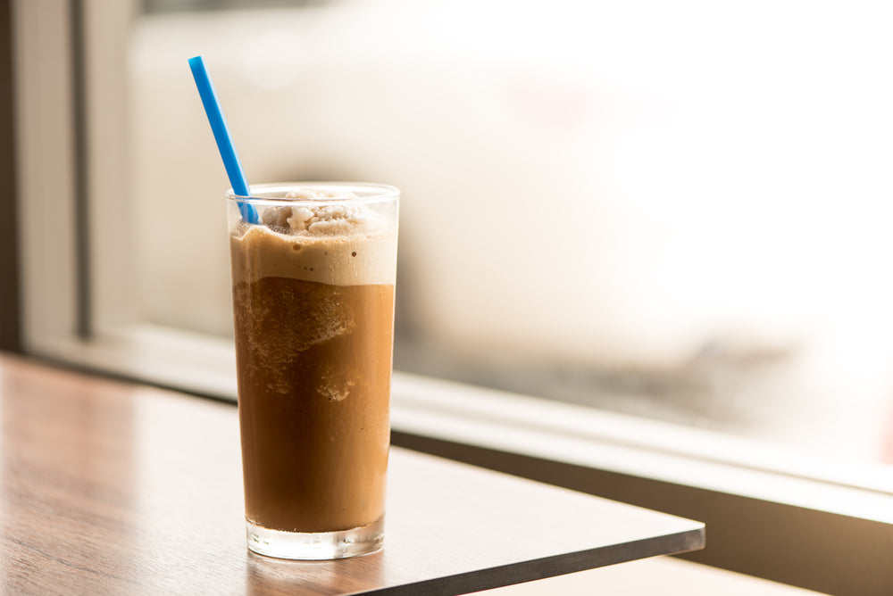 A tall glass filled with a creamy, frothy French vanilla cola float sits on a wooden table, featuring a blue straw and a soft-focus background of a bright window.