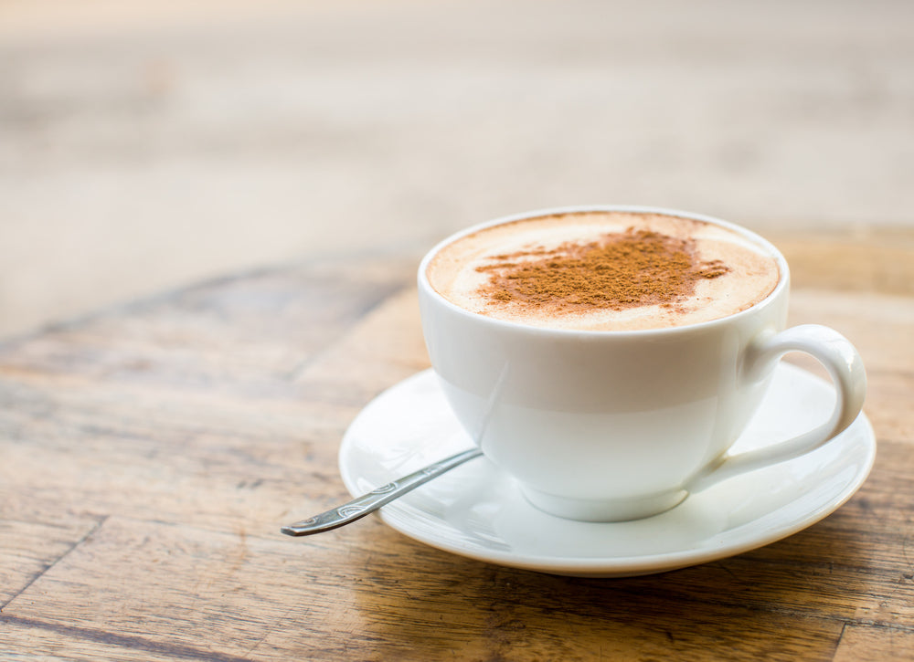 A white cup filled with a creamy vanilla cappuccino smoothie, topped with a sprinkle of cinnamon, sits on a round white saucer on a wooden table, with a silver spoon resting beside it.