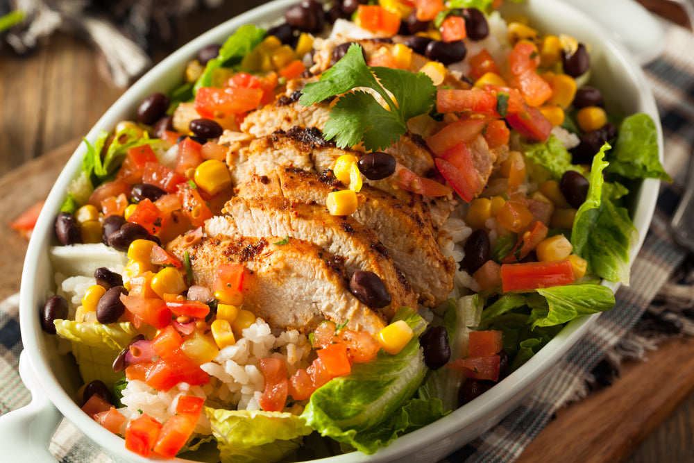 A bowl of Mexican chicken salad features sliced grilled chicken on a bed of lettuce, topped with corn, black beans, diced tomatoes, and cilantro, with a rustic wooden background.
