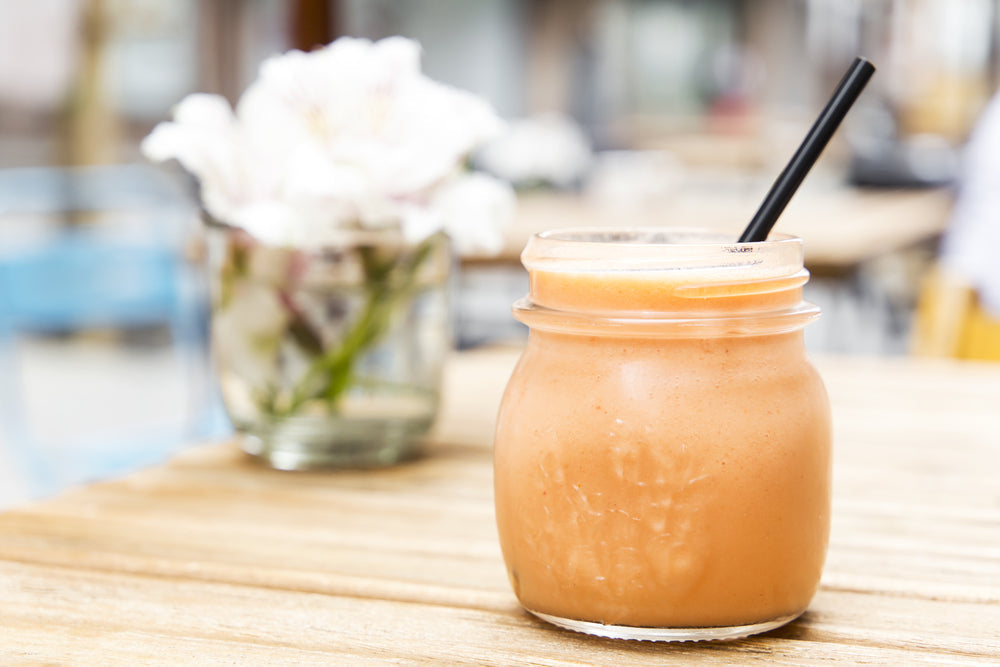 A creamy fruity shake in a glass jar with a black straw sits on a wooden table, next to a vase of white flowers, in a bright outdoor café setting.