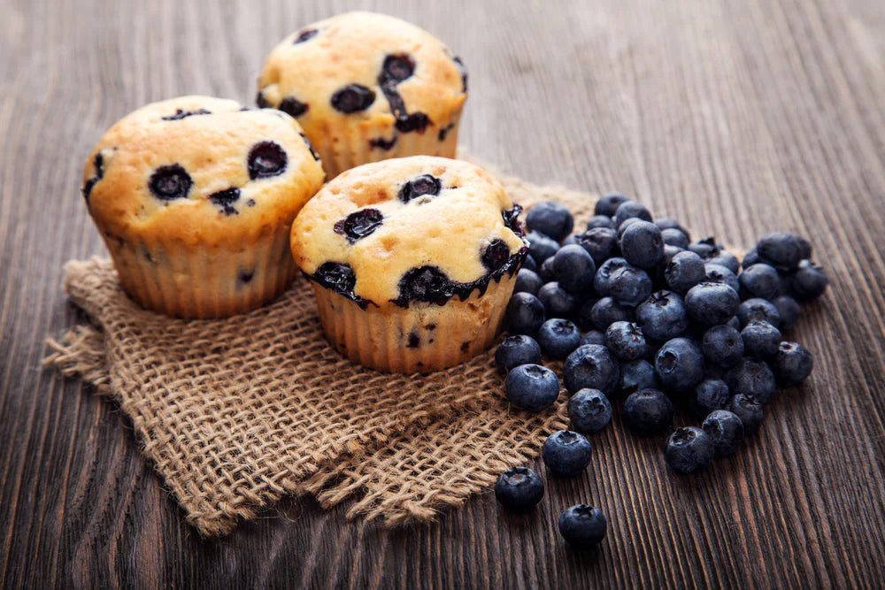 Three freshly baked blueberry muffins sit on a burlap cloth beside a pile of blueberries, all arranged on a wooden surface.