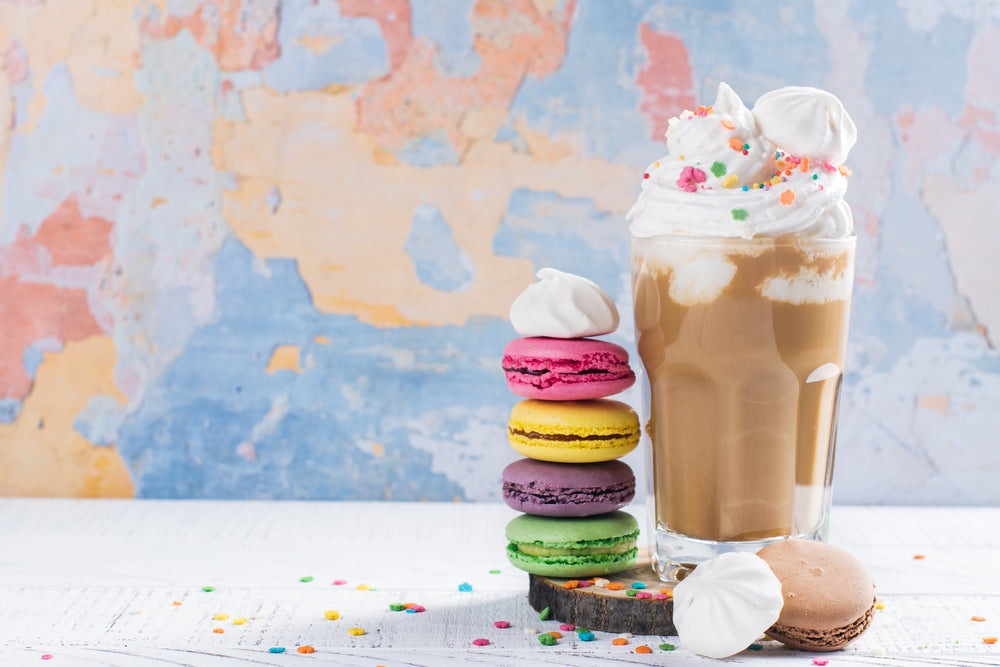 A tall glass of amaretto cookie shake topped with whipped cream and colorful sprinkles sits beside a stack of five macarons in various colors against a softly textured pastel background.