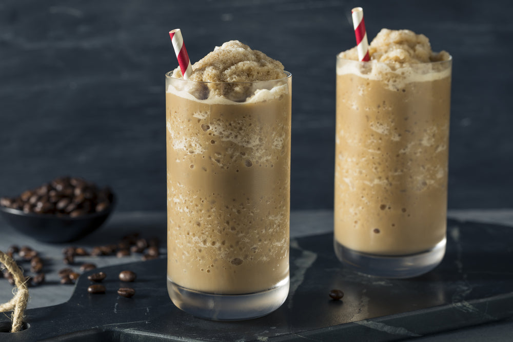 Two tall glasses of frozen latte topped with whipped cream and red-and-white striped straws sit on a dark surface, with a small bowl of coffee beans nearby.