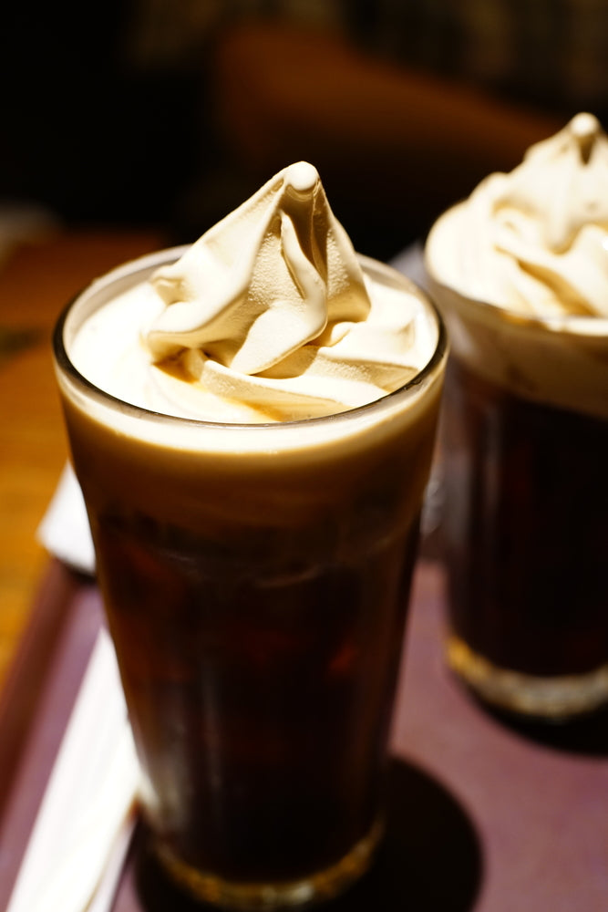 Two tall glasses of root beer float topped with creamy, swirled chocolate-flavored foam are placed on a dark tray, with a blurred background suggesting a cozy dining setting.