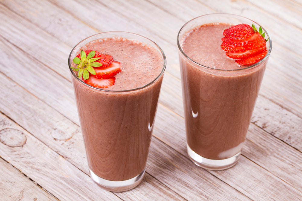 Two tall glasses of chocolate-strawberry smoothie sit on a wooden table, each topped with a slice of fresh strawberry and a small green leaf, showcasing a delicious and refreshing drink.