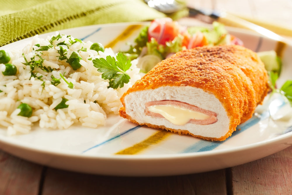 A plate of Chicken Cordon Bleu, featuring a golden-breaded chicken breast stuffed with ham and cheese, served alongside fluffy rice with peas and a fresh salad.