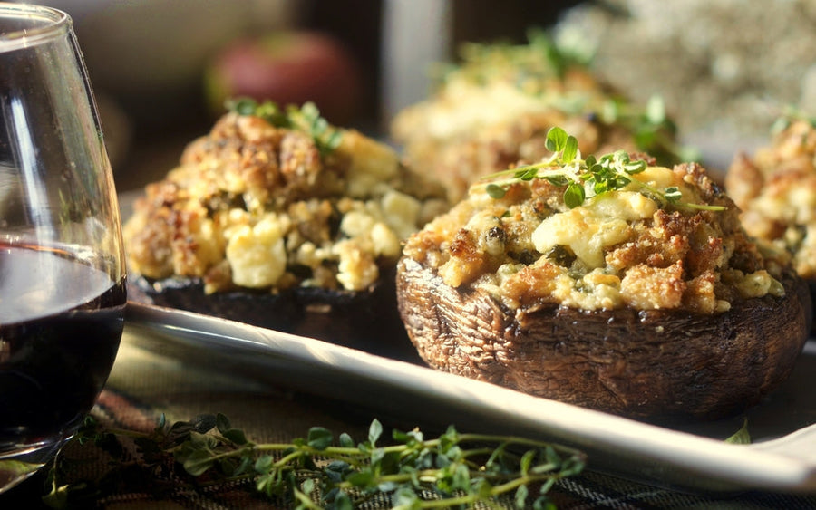 Three stuffed mushrooms topped with herbs sit on a platter next to a glass of red wine, with a blurred background suggesting a cozy dining setting.