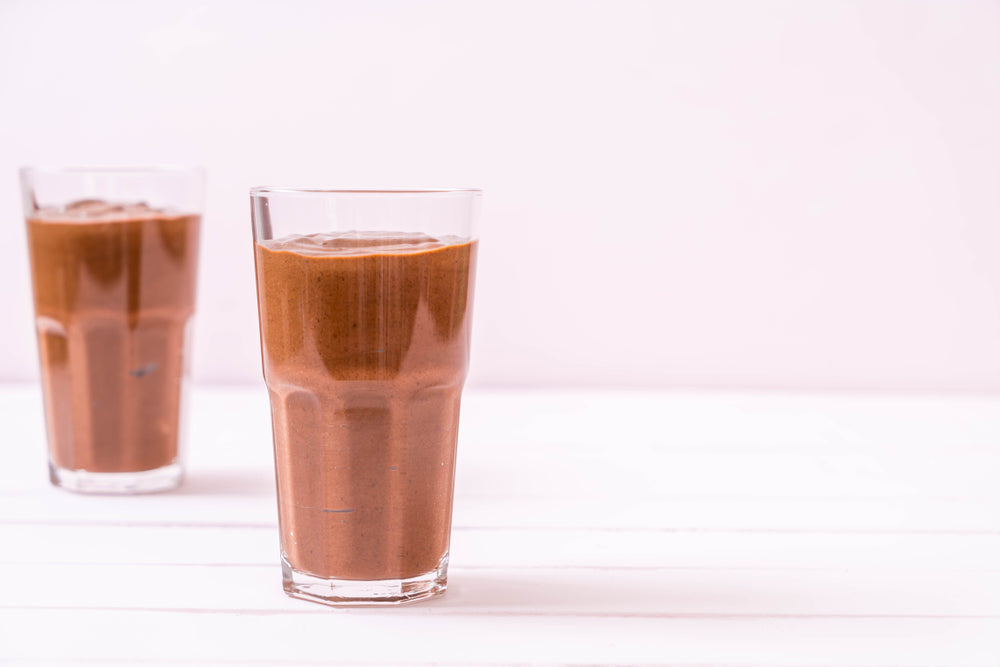 Two tall glasses filled with a creamy chocolate beverage sit on a white surface, with one glass slightly behind the other, against a soft pink background.