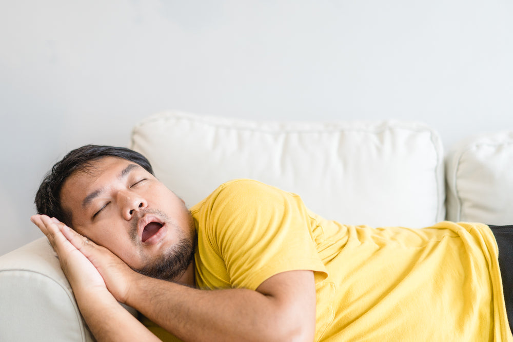 A man in a yellow shirt is sleeping on a white couch, resting his head on his hands with his mouth slightly open, illustrating a moment of rest that may relate to sleep apnea.